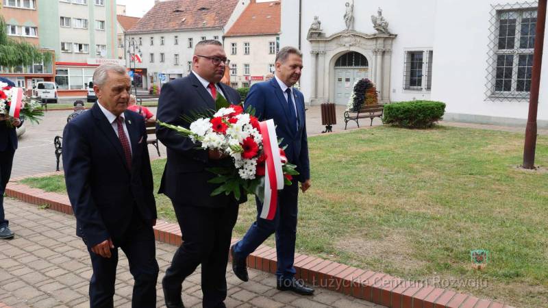 trzy osoby idą, od lewej: Stanisław Jastrzebski, Kamil glazer (niesie biało-czerwone kwiaty), Arkadiusz Słowiński